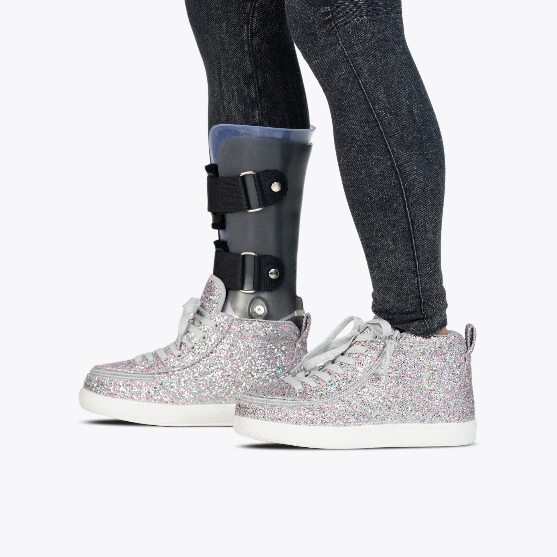A person wears Silver Multi Glitter BILLY Classic D|R II High sneakers by BILLY Footwear, paired with dark jeans and a visible left leg prosthetic brace. The orthotic-friendly shoes are shown against a plain white background.