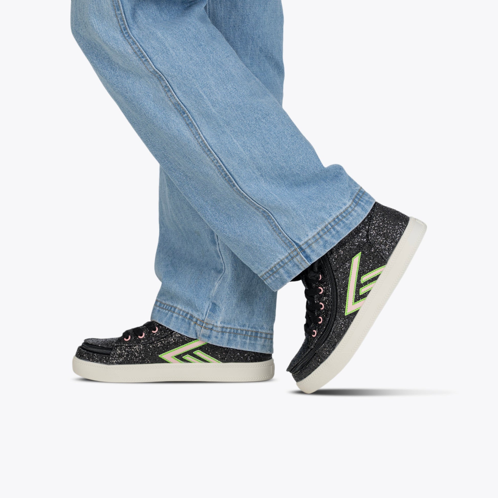 A person in light blue jeans and Wicked Black Glitz BILLY CS High sneakers by BILLY Footwear with green and white chevron designs walks against a plain white background.