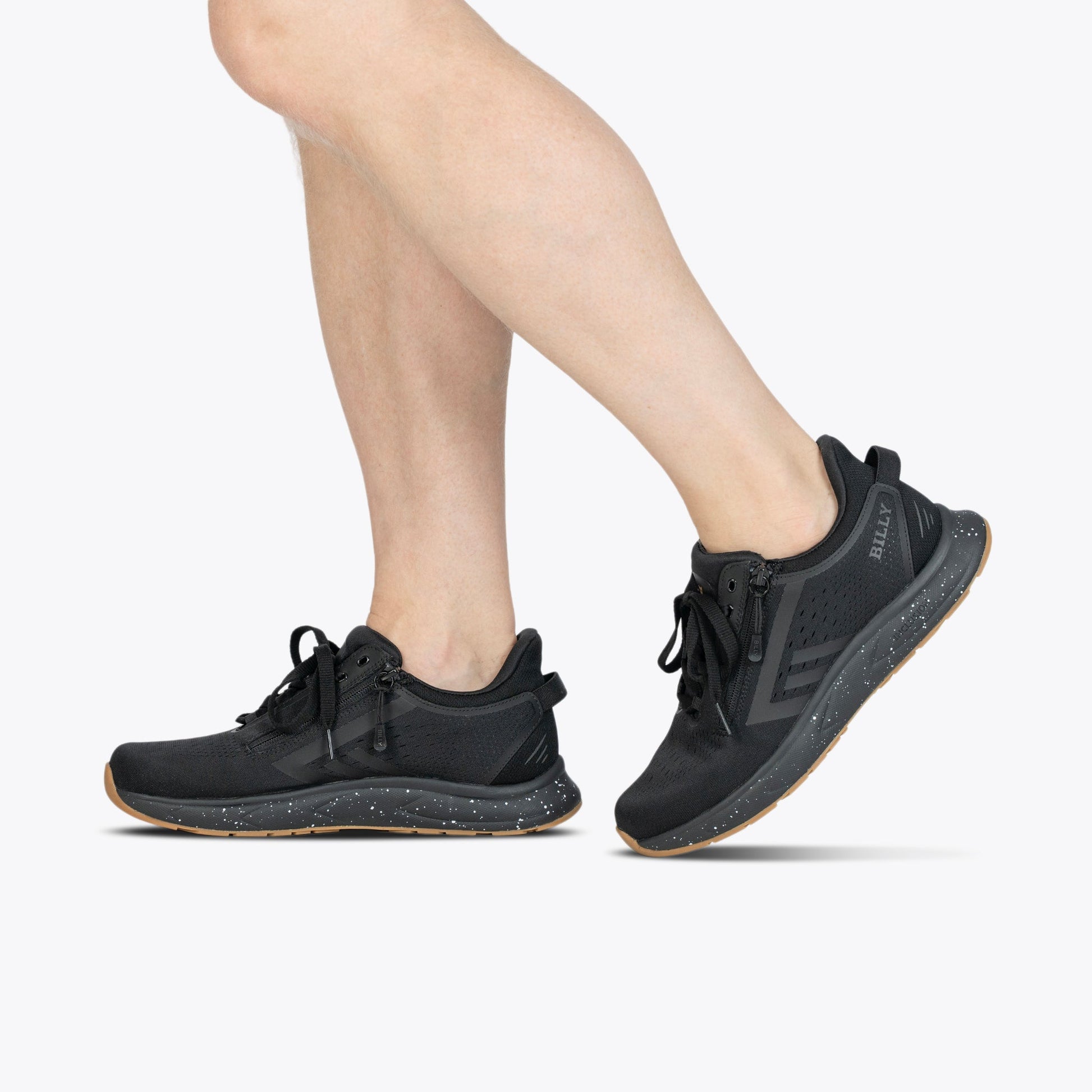 A person wears BILLY Footwear Men's Black/Black/Gum BILLY Sport Inclusion DZ1 sneakers, standing with one foot slightly ahead of the other against a plain white background.