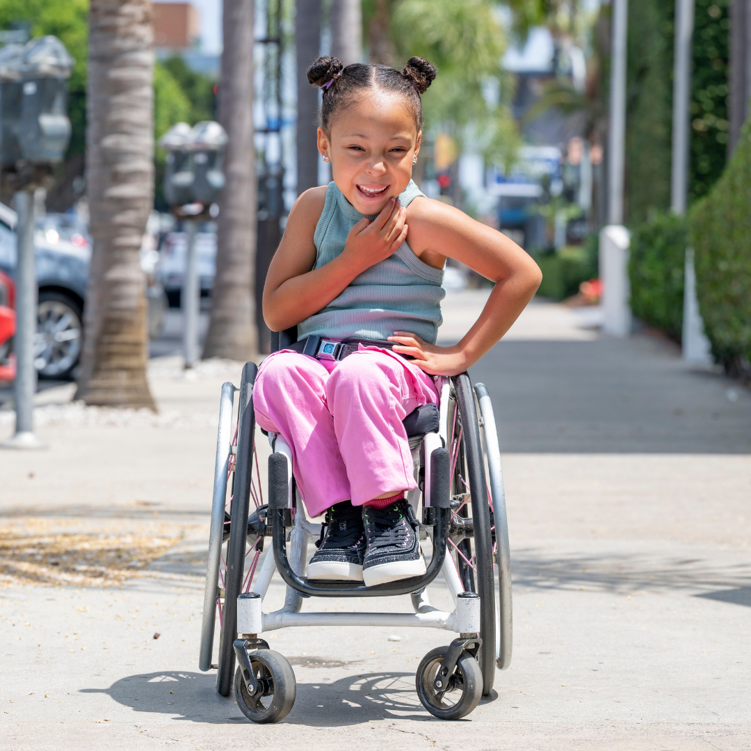 A young girl in a wheelchair smiles on a sunny sidewalk, wearing a striped tank top, pink pants, and Wicked Black Glitz BILLY CS High shoes by BILLY Footwear. Palm trees and parked cars fill the background as her hand rests on her chest.
