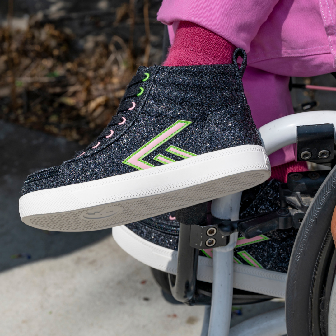 A person in pink pants rests their foot on a wheelchair footrest outdoors while wearing Wicked Black Glitz BILLY CS High shoes by BILLY Footwear, featuring green and pink accents.